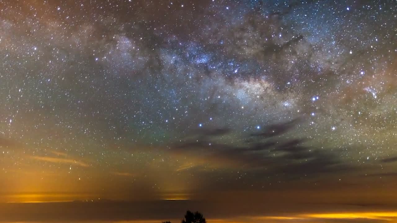 Imágenes en time-lapse de un cielo nocturno estrellado con la galaxia Vía Láctea extendiéndose por el horizonte. Innumerables estrellas brillan sobre árboles silueteados, mientras que tonos atmosféricos de verde, naranja