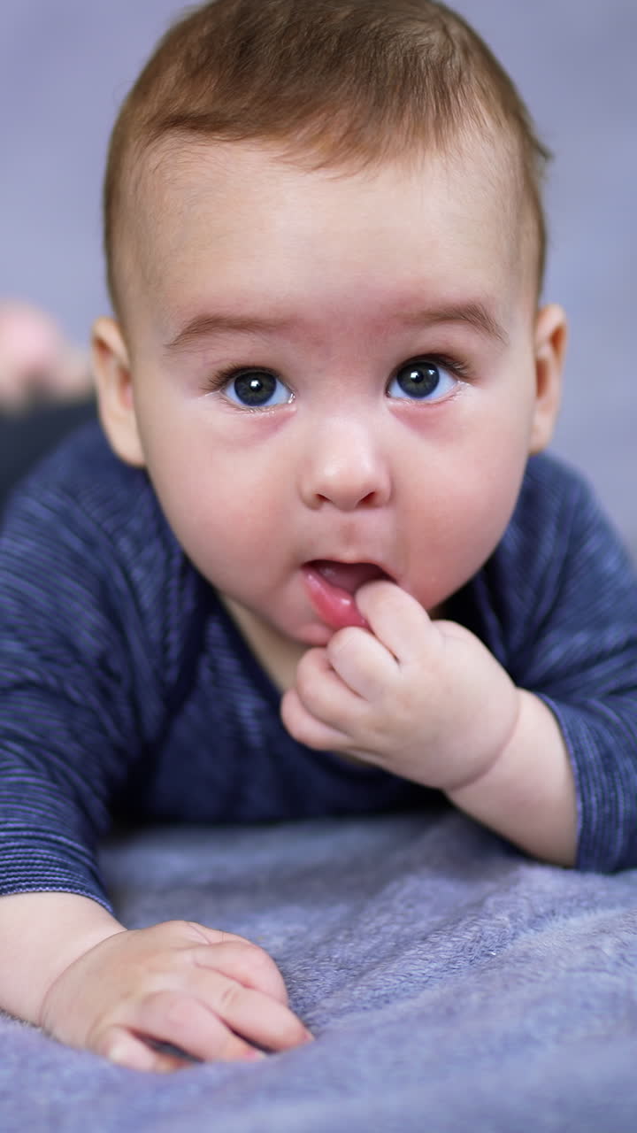 Adorable Caucasian child lies on belly with his finger in mouth. Lovely sweet baby resting peacefully on the grey plaid. Close up. Blurred backdrop. Vertical video