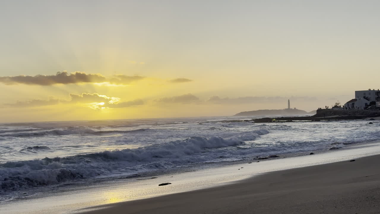 Sun setting over gentle waves with a silhouette of a lighthouse and coastal buildings