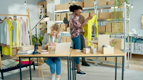 caucasian designer man typing on tablet and checking boxes while talking with female designer coworker in fashion clothing shop