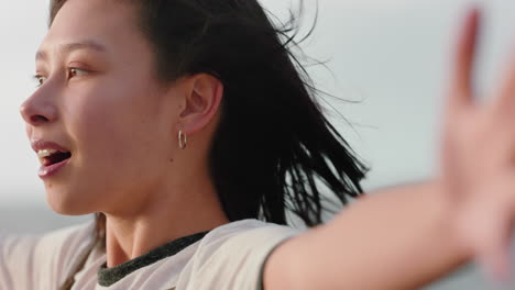 portrait of happy asian woman enjoying freedom arms raised feeling joy on beach at sunset exploring wanderlust with wind blowing hair