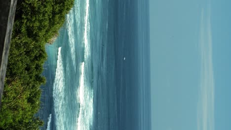 Vertical-Static-shot-of-a-limestone-cliff-face-overlooking-waves-rolling-in-to-an-island-with-a-handrail-in-the-foreground,-and-green-brush