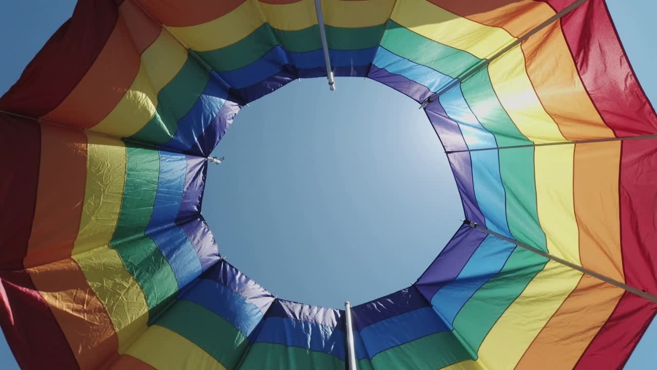 Ombrellone arcobaleno ondeggiante che cattura il sole in spiaggia a causa di una leggera brezza, rivelando il palo metallico e il cielo.