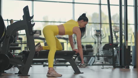 Hispanic-athlete-woman-in-sportswear-training-back-with-dumbbell-in-one-hand-leaning-on-the-bench-at-fitness-gym