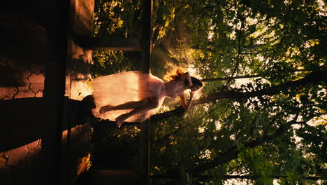 Beautiful-Woman-Sitting-on-Bridge,-Backlit-By-Beautiful-Golden-Sunlight,-Dream-Scene,-Sun-flares,-Forest-Background,-Cinematic-Silhouette,-Golden-Hour,-Slow-Motion-Vertical,-Camera-Tracks-Back