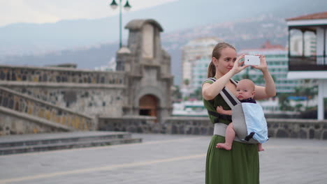 carrying her infant in a kangaroo backpack, a young woman takes snapshots on a mobile phone during her travels. she walks and periodically gazes at the phone screen