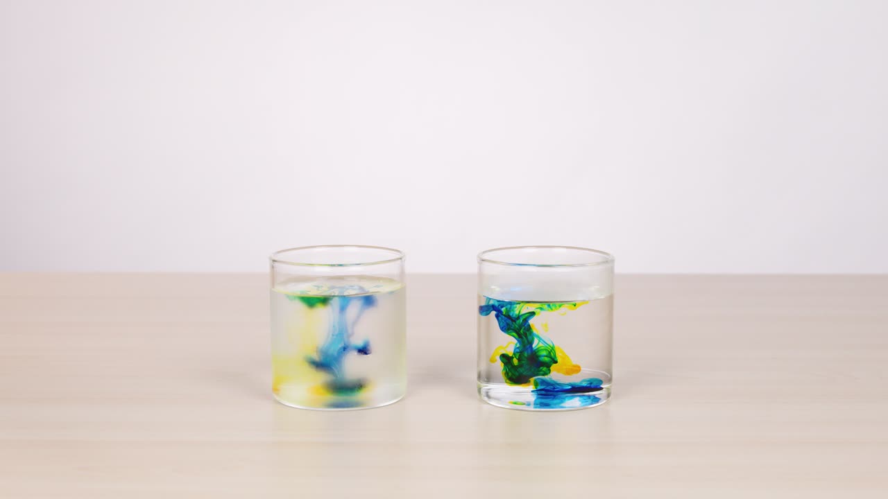 Colored water droplets disperse in two clear glasses under bright, even lighting on white background