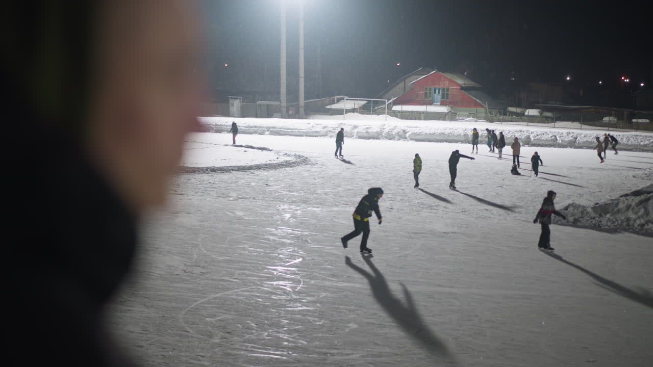 Blurred figure in foreground watches group of people skating on illuminated outdoor ice rink surrounded by snow, as shadows stretch across frozen surface under powerful night lighting