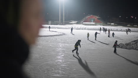 blurred figure in foreground watches group of people skating on illuminated outdoor ice rink surrounded by snow, as shadows stretch across frozen surface under powerful night lighting