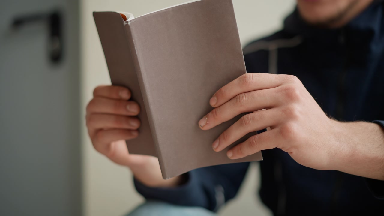 Primo piano di un uomo che tiene e legge un libro aperto con entrambe le mani. L'uomo indossa abiti casual, concentrandosi sull'interazione tra le sue dita e la copertina testurizzata di un taccuino con copertina rigida. Illuminazione soffusa.