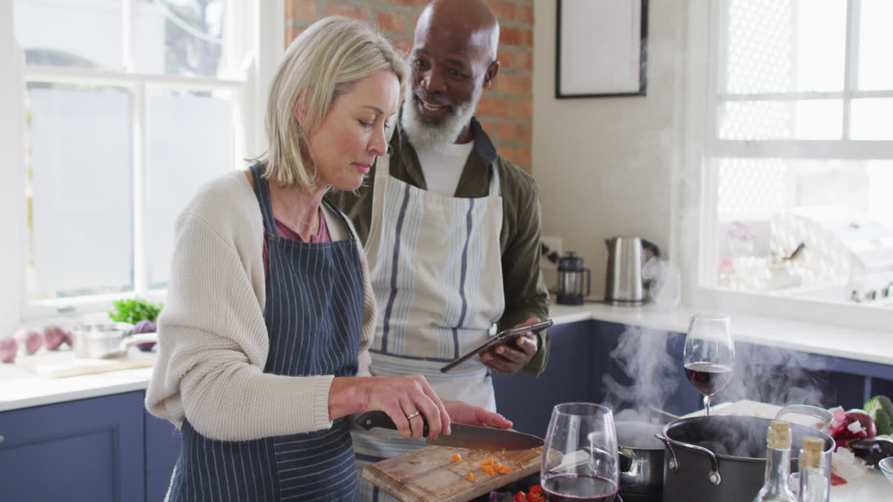 pareja de adultos de raza mixta usando delantales usando una tableta digital mientras cocinan en la cocina en casa