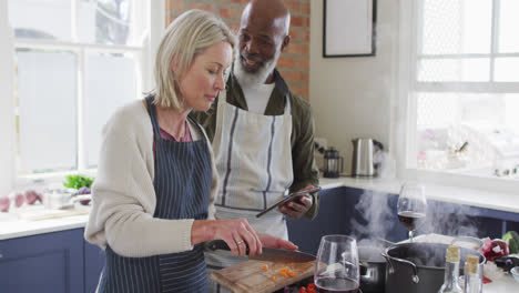 Pareja-De-Ancianos-De-Raza-Mixta-Usando-Delantales-Usando-Una-Tableta-Digital-Mientras-Cocina-En-La-Cocina-De-Casa