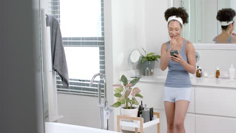 mujer biracial feliz cepillándose los dientes y usando un teléfono inteligente en el baño, cámara lenta