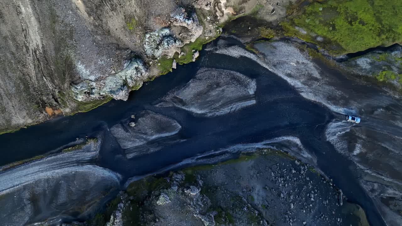 Drone view of an expedition SUV following a braided volcanic river in Iceland’s rugged highlands, capturing dark sediment channels, eroded slopes, and remote wilderness terrain from above