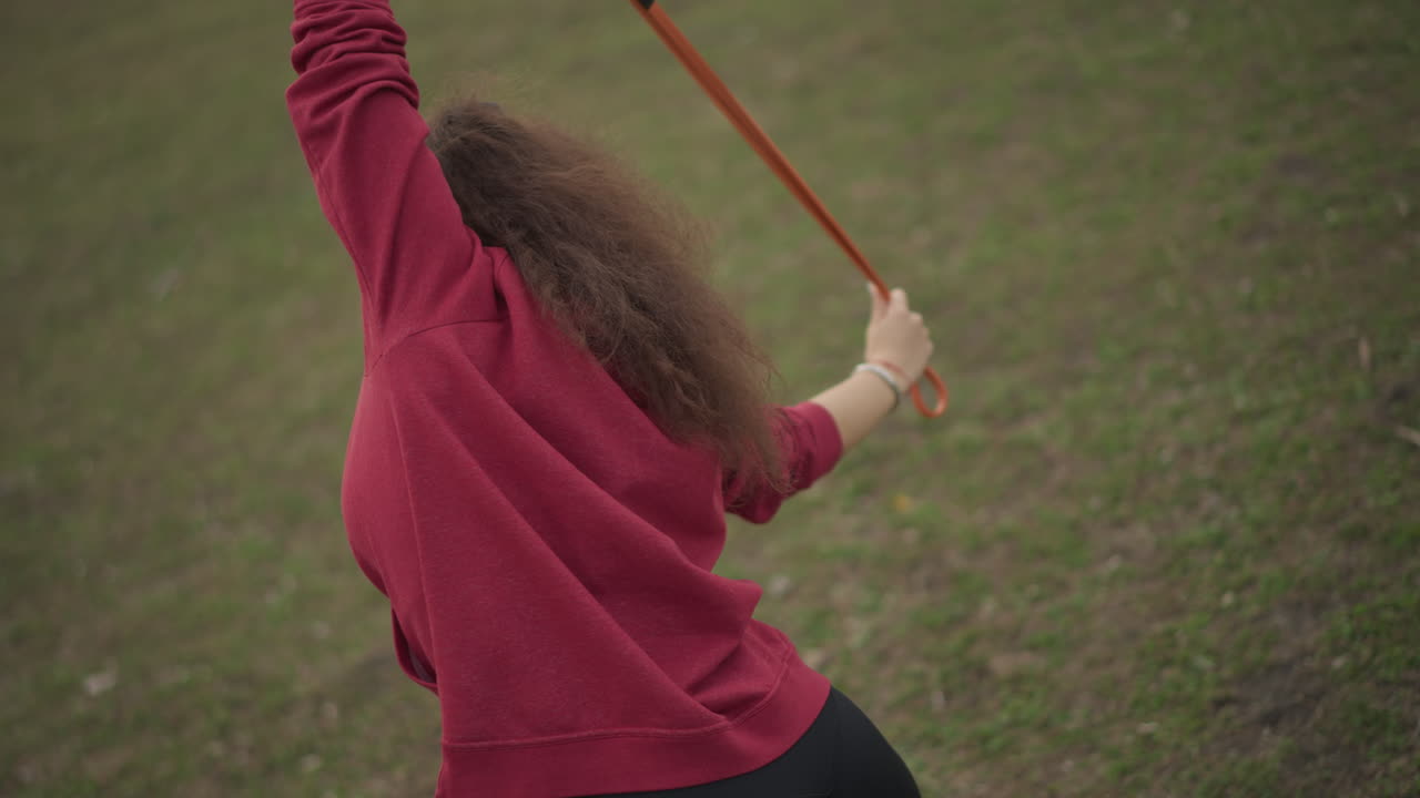 Energetic White Woman Swinging Orange Band Outdoors, Athlete Performing Dynamic Resistance Drills Across Grassy Park, Focused Posture And Active Training Vibe With Hoodie And Ponytail