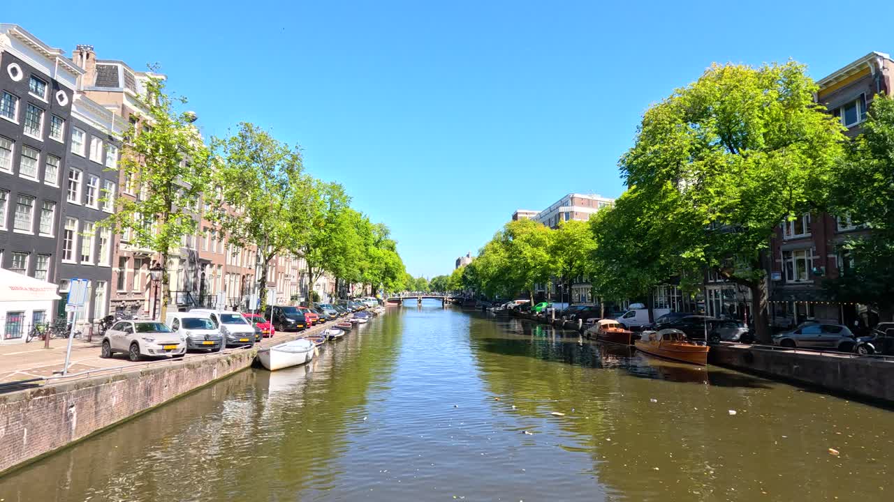 Wide shot of Amsterdam canal, boats, historic buildings, and trees on a bright summer day