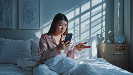 Confused woman in pajamas looking at her phone in bed