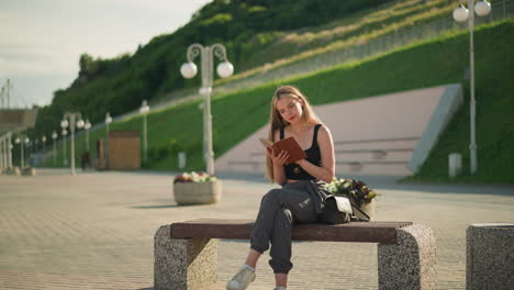 woman seated outdoors on bench, reading a book thoughtfully with legs crossed, sun illuminating her, she holds the book gently, while the background features lined-up light poles