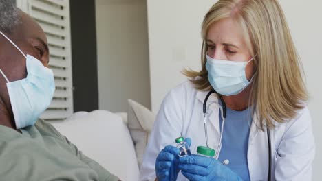 Doctora-Caucásica-De-Alto-Rango-Mostrando-Medicación-A-Un-Paciente-Masculino-En-La-Cama,-Ambos-Usando-Máscaras-Faciales