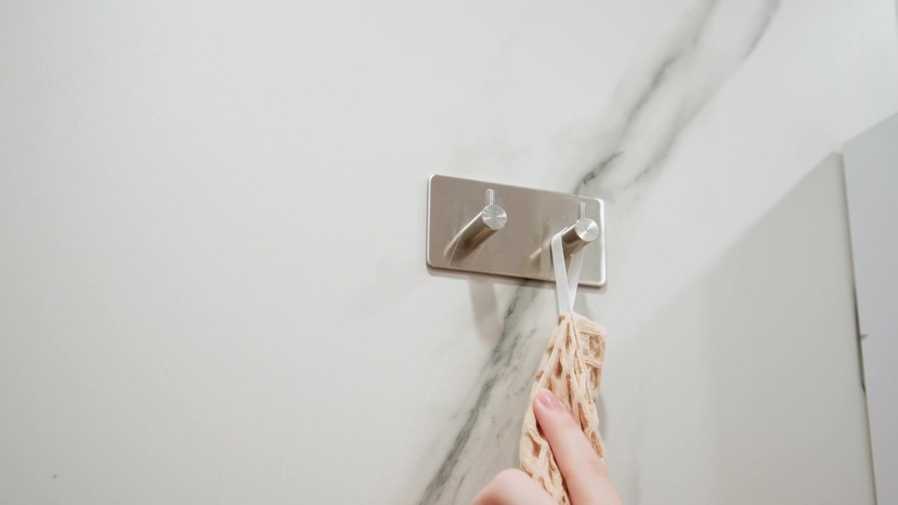 Hand placing woven cloth onto metal double-prong wall hook beside marbled white tile wall with gray veining, glossy cabinet edge, soft reflections, and bright indoor bathroom lighting