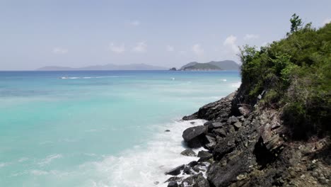 Low-aerial-view-of-blue-water-crashing-on-rocks-USVI-virgin-islands-boats-crossing-scene-blue-sky-white-clouds-tranquil-relaxation-mountains-in-background