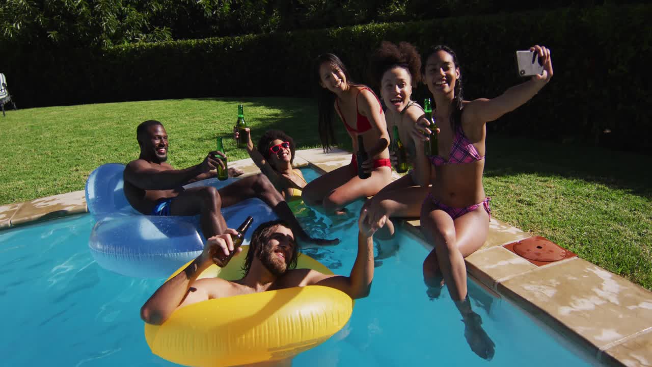 Gruppo eterogeneo di amici seduti a bordo piscina che bevono birra e si fanno un selfie.