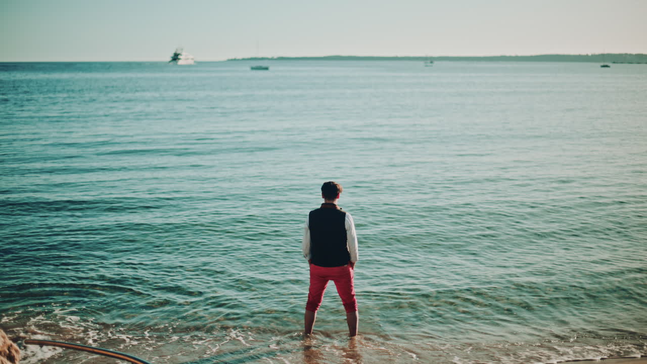 A man standing at the shoreline, facing the sea as small waves reach his feet