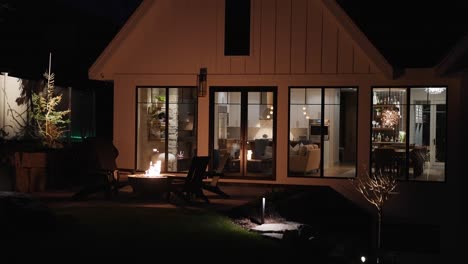 Exterior-home-at-night-with-firepit,-flickering-firelight-on-house,-dimly-lit-living-room-visible