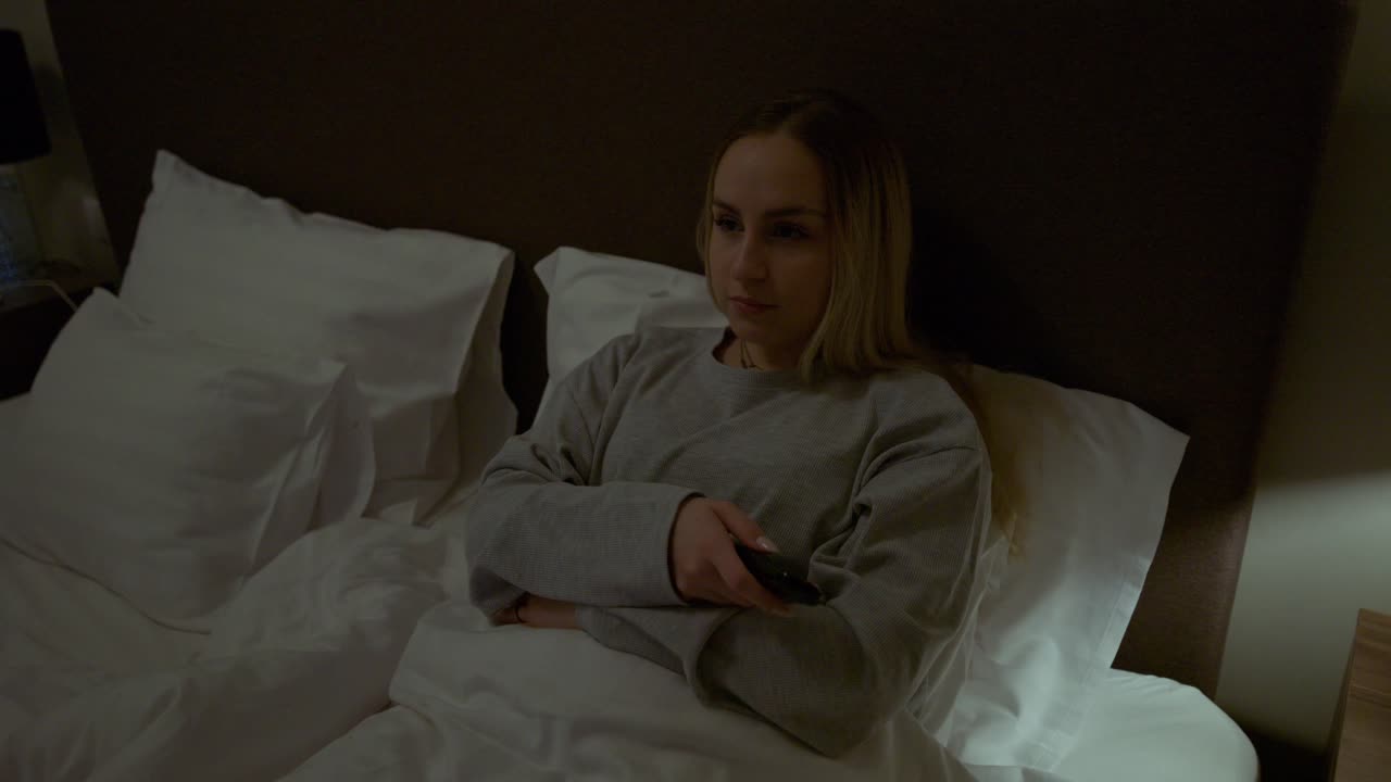 Una giovane donna comodamente sdraiata a letto punta un telecomando verso la televisione in una camera da letto illuminata da luce soffusa. La calda lampada da comodino e la biancheria da letto accogliente creano un'atmosfera domestica serale rilassata.