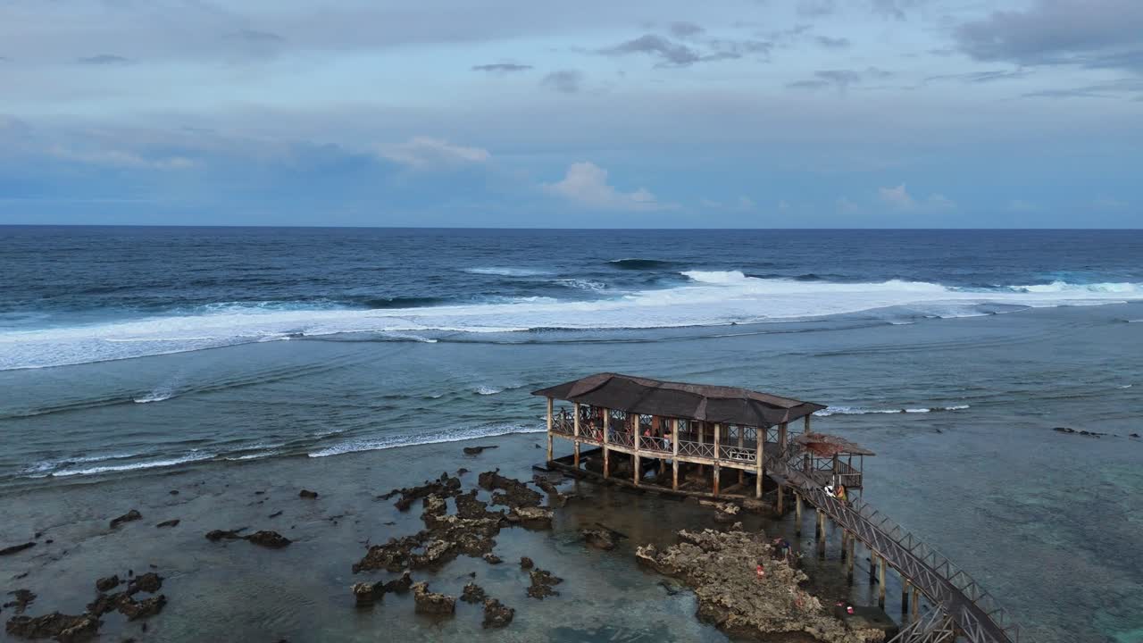 Drone orbits above famous Cloud 9 Surfing Area in Siargao, Philippines, showing wooden pier and observation deck extending over shallow reef and surf waves rolling in from the Pacific Ocean