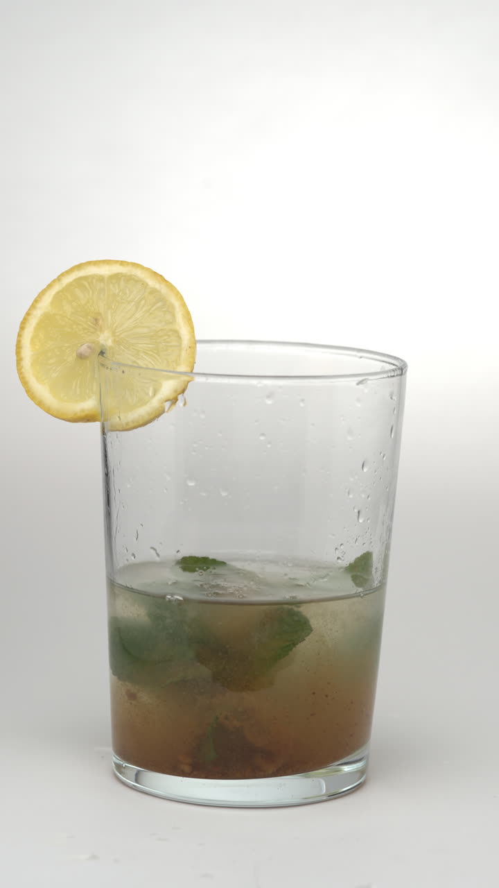 Video verticale 9:16 al rallentatore di un cucchiaio che versa zucchero moscovado in un bicchiere di limonata con menta e una fetta di limone su uno sfondo bianco pulito