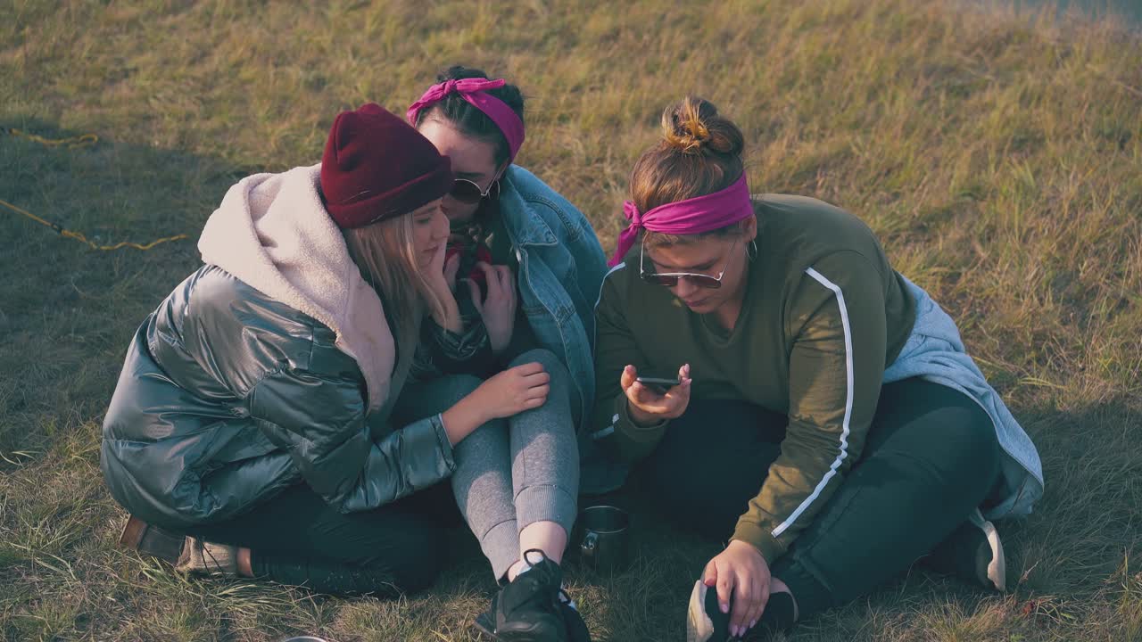 Kadın turistler çimlerde oturarak akıllı telefonda video izliyor.