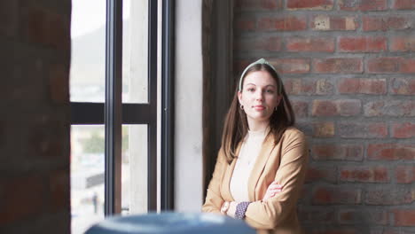 Young-Caucasian-businesswoman-leans-against-window,-arms-crossed,-with-a-subtle-smile