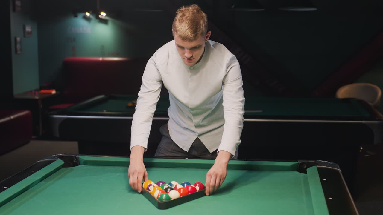 deportista con camisa blanca y pantalones grises arregla cuidadosamente bolas de billar de colores en formación de triángulo en una mesa de billar verde vibrante, en una elegante sala de billar con otra mesa visible
