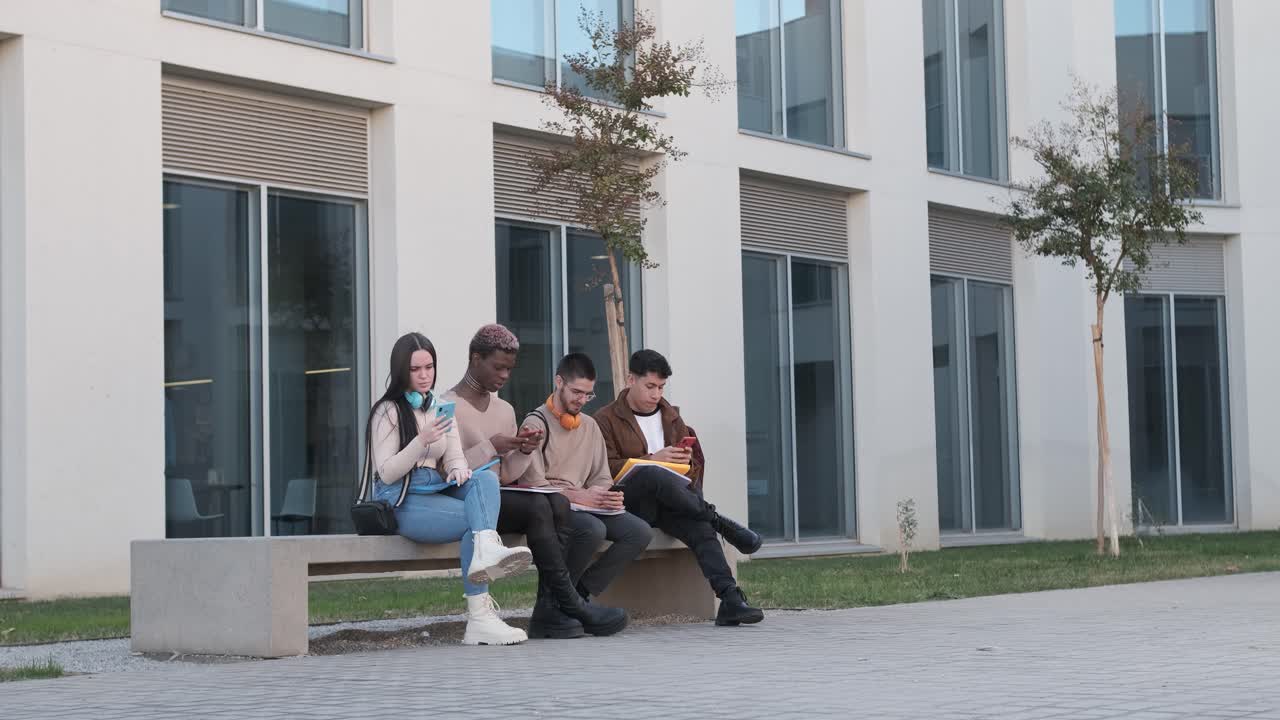 Mulitcultural students using their own mobile sitting in a bench outdoors