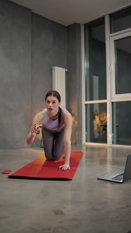 vertical video of a confident brunette girl in a sports summer uniform doing sports on a red sports mat using video lessons via a laptop in a modern apartment