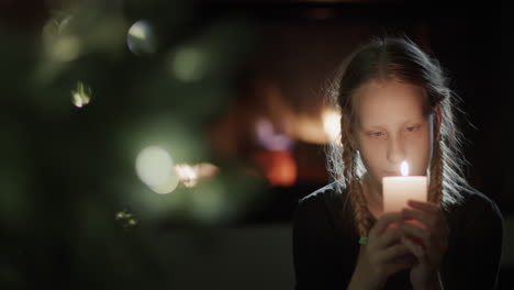 a teenage girl holds a burning candle in her hands. sits by the christmas tree in the background a fire is burning in the fireplace. make a wish on christmas eve