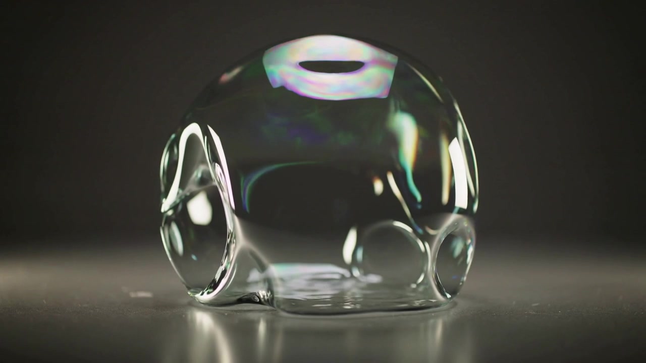 Deforming single clear soap bubble flattening on studio table as film thins, leaving wet patch