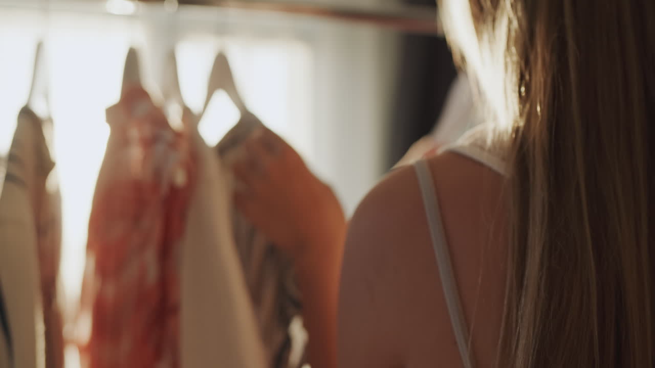 십대 소녀는 무엇을 입을지 선택합니다. 옷은 그녀의 앞에서 어깨에 매달려 있습니다. 태양은 옷을 조명합니다.