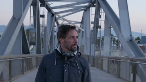 Front-view-casual-short-bearded-male-walking-across-breezy-iron-girder-bridge