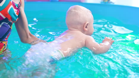 Beautiful blond Caucasian baby wearing a diaper is taught to swim. Female coach splashes water on the back of a child.
