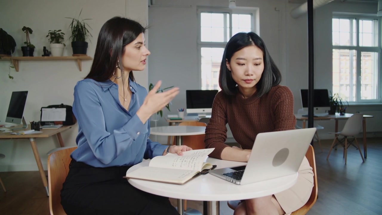 オフィスで、青いブラウスを着た人物がノートを開き、ノートパソコンを使いながら、同僚にプロジェクトを指導している。