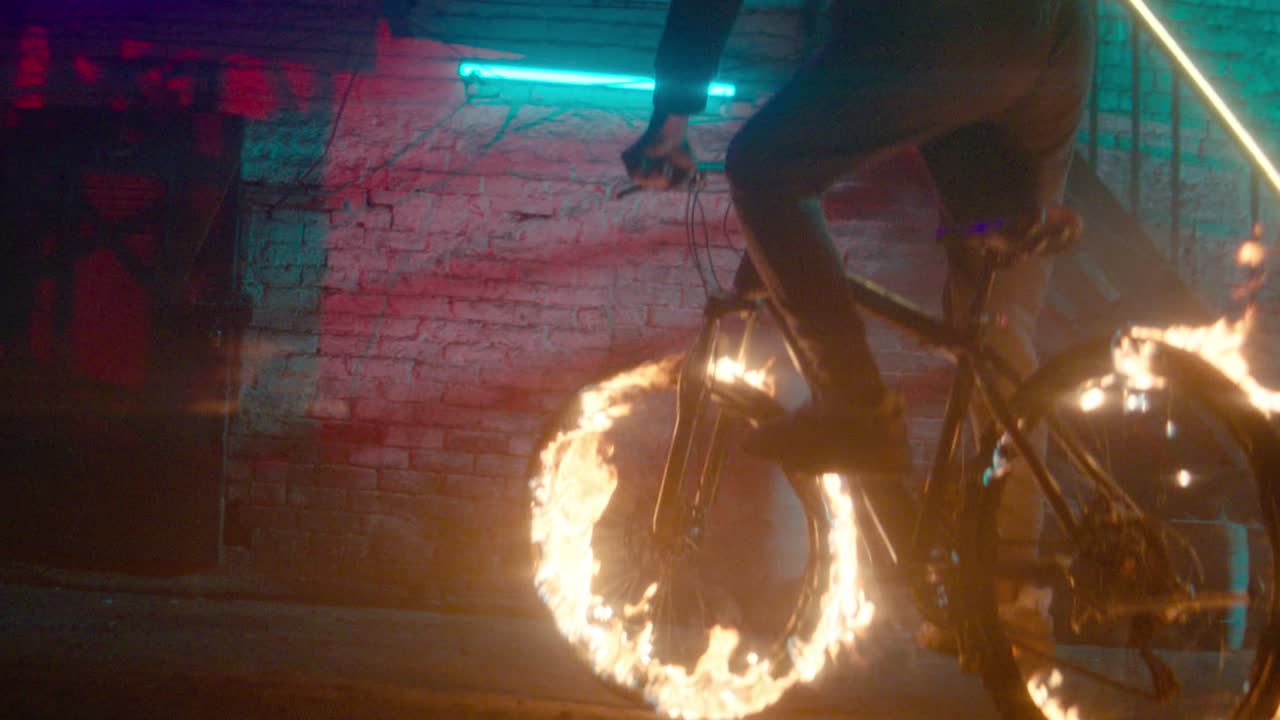 Metraje cinematográfico en súper cámara lenta de un ciclista pedaleando por un entorno urbano con ambas ruedas de la bicicleta envueltas en llamas. Iluminación de neón roja y azul que se refleja en una pared de ladrillo y metal