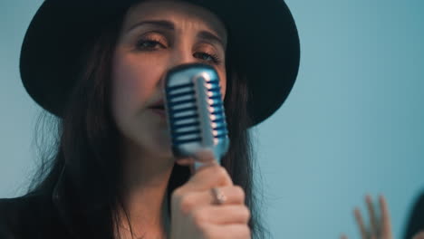 a singer in a black hat and black leather jacket holds a vintage microphone with one hand while passionately gesturing with the other, set against a blue background