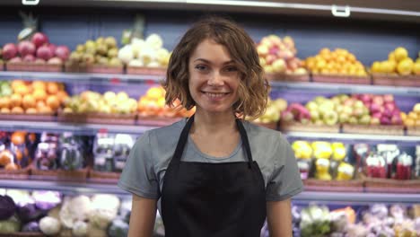 retrato de una joven vendedora atractiva con un delantal negro de pie en un supermercado con estantes de frutas en el fondo, mirando a la cámara y sonriendo. concepto de negocios comerciales y personas