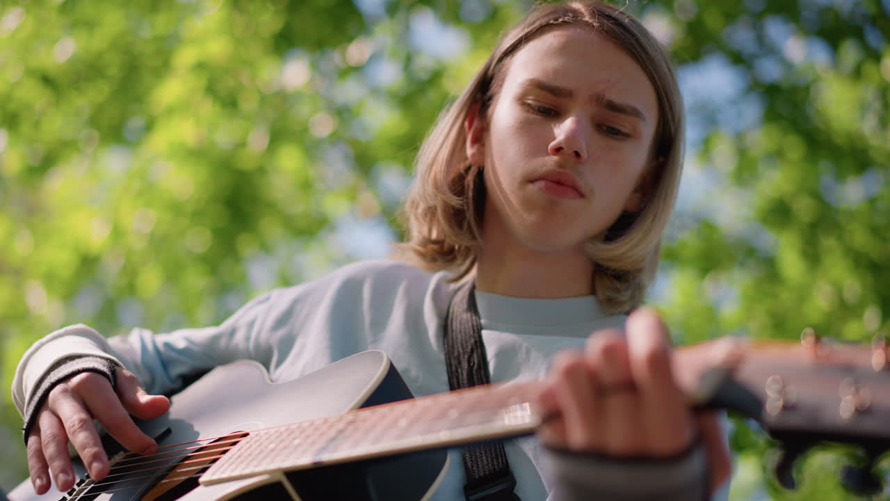 giovane chitarrista bianco che suona la chitarra acustica in un parco illuminato dal sole, ritratto dal basso che mostra un'espressione concentrata, dita sulla tastiera, sfondo bokeh verde, maglione casual, atmosfera indie folk, pacifico
