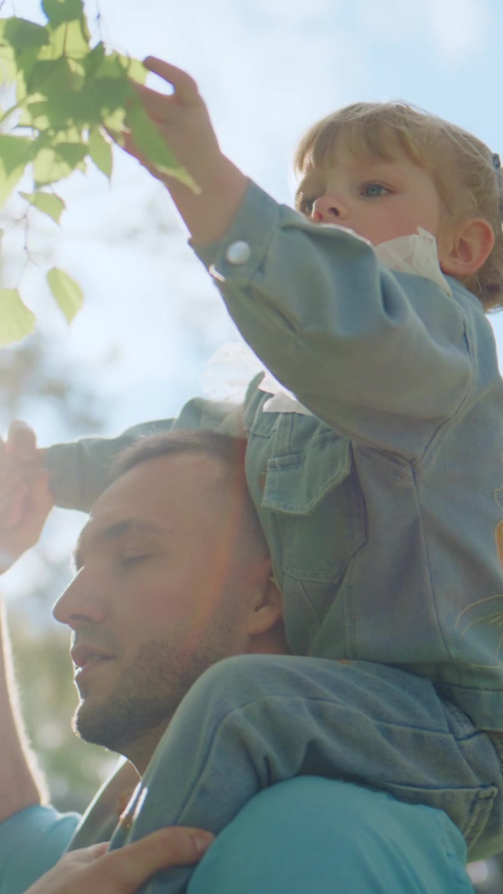 Il padre porta il figlio sulle spalle all'aperto. Il bambino si protende verso le foglie, curioso e deliziato. Il padre guarda in alto, sorridendo dolcemente con orgoglio. La scena cattura un gioioso momento di legame familiare.