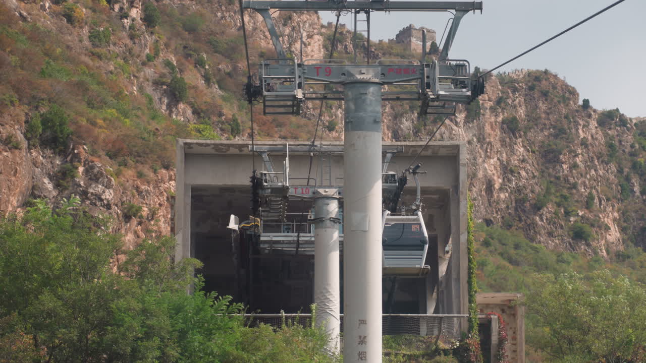 Bergseilbahn fährt in die Station ein. Betonterminal umrahmt von felsigen Klippen und dichtem Gestrüpp. Hoher Stahlpylon im Vordergrund. Gondelkabinen gleiten auf straffen Seilen in den dunklen Tunneleingang.