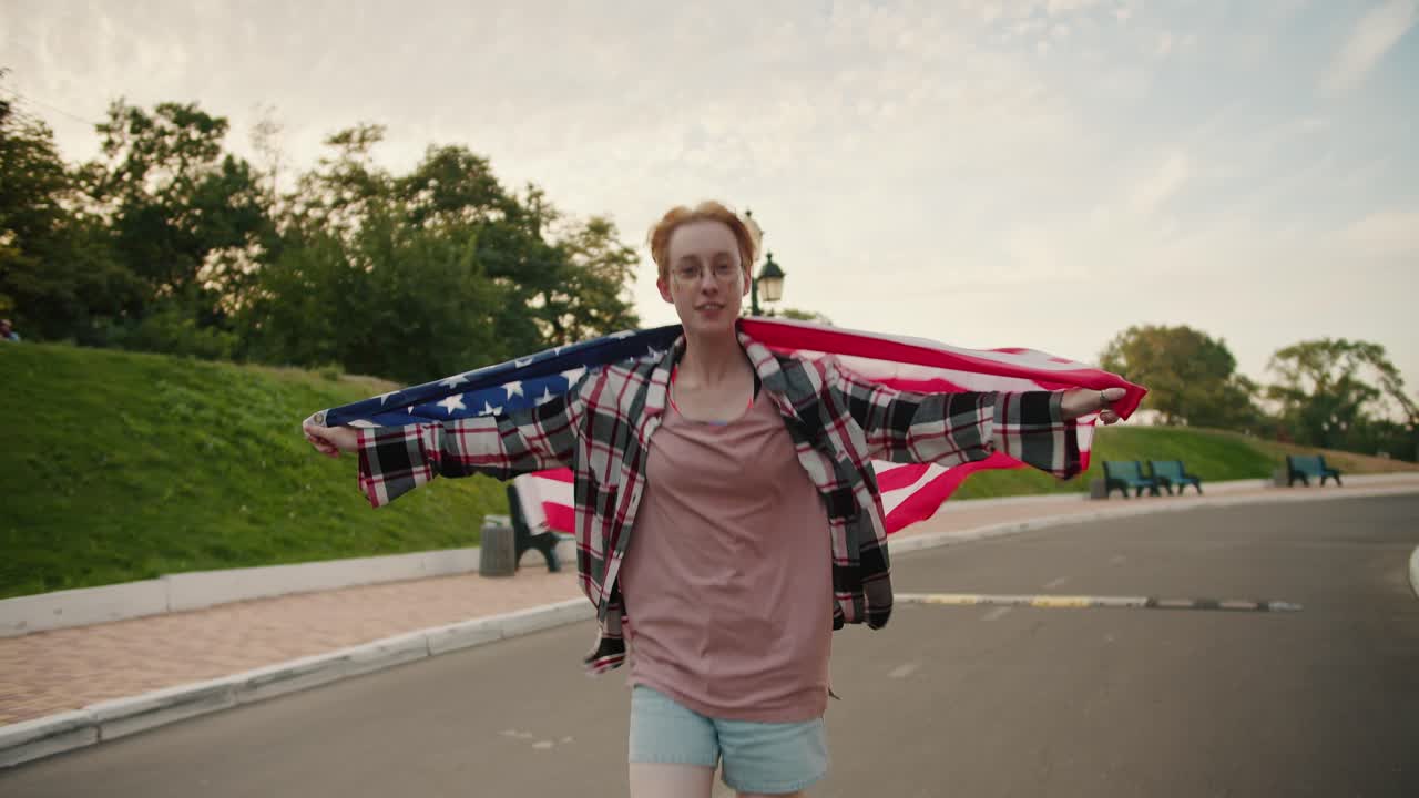A blonde girl with short hair with glasses in a checkered pink shirt runs with the flag of the United States of America behind her shoulders along the road in the park in the summer