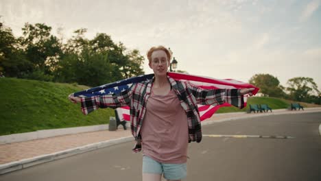 A-blonde-girl-with-short-hair-with-glasses-in-a-checkered-pink-shirt-runs-with-the-flag-of-the-United-States-of-America-behind-her-shoulders-along-the-road-in-the-park-in-the-summer